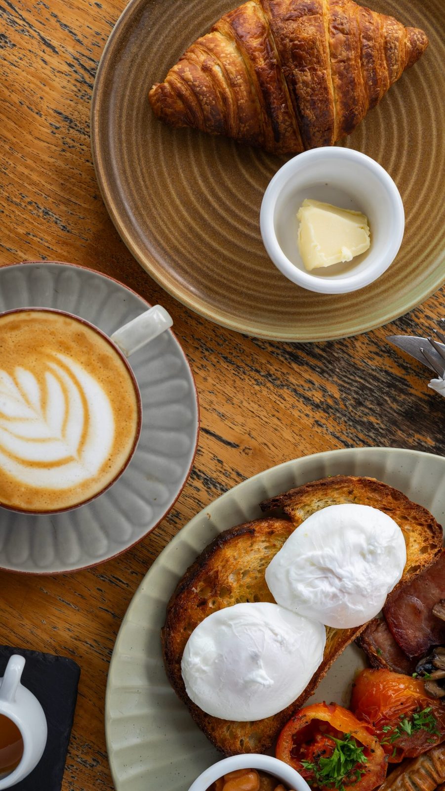 Flat white, croissant with butter and poached eggs on sourdough on a teak table