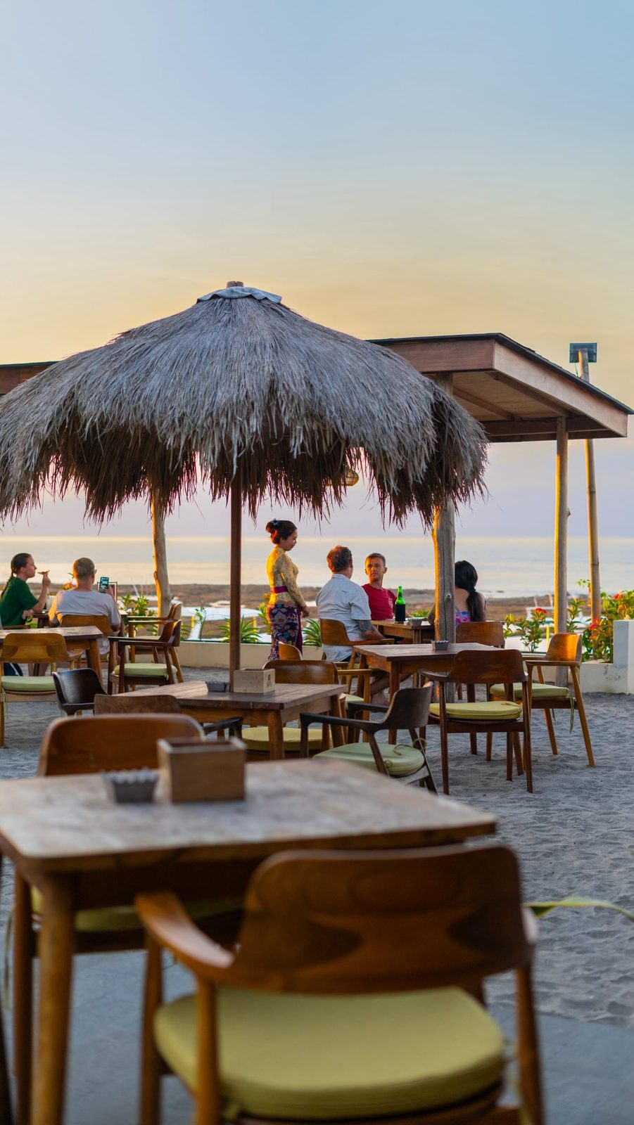 Thatched umbrella over teak tables at sunset, guests at the water's edge